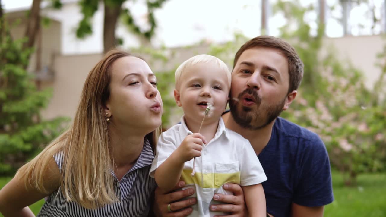 familia joven jugando en el parque. padre, madre y bebé niño feliz se sentó juntos cerca de la flor. niño rubio con flores, diente de león en las manos y soplando en ellos. mamá y papá se unen a su hijo en el soplo. familia feliz. retrato. cámara lenta