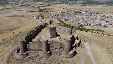 Vuelo-Vital-En-Un-Castillo-En-Ruinas-Con-Sus-Murallas-Y-Torres-En-Una-Colina-Viendo-Que-La-Torre-Principal-Está-En-Medio-De-La-Fortaleza-Y-Con-Campos-De-Cultivo-Y-Un-Pueblo-En-Un-Día-De-Verano-En-Toledo,-España