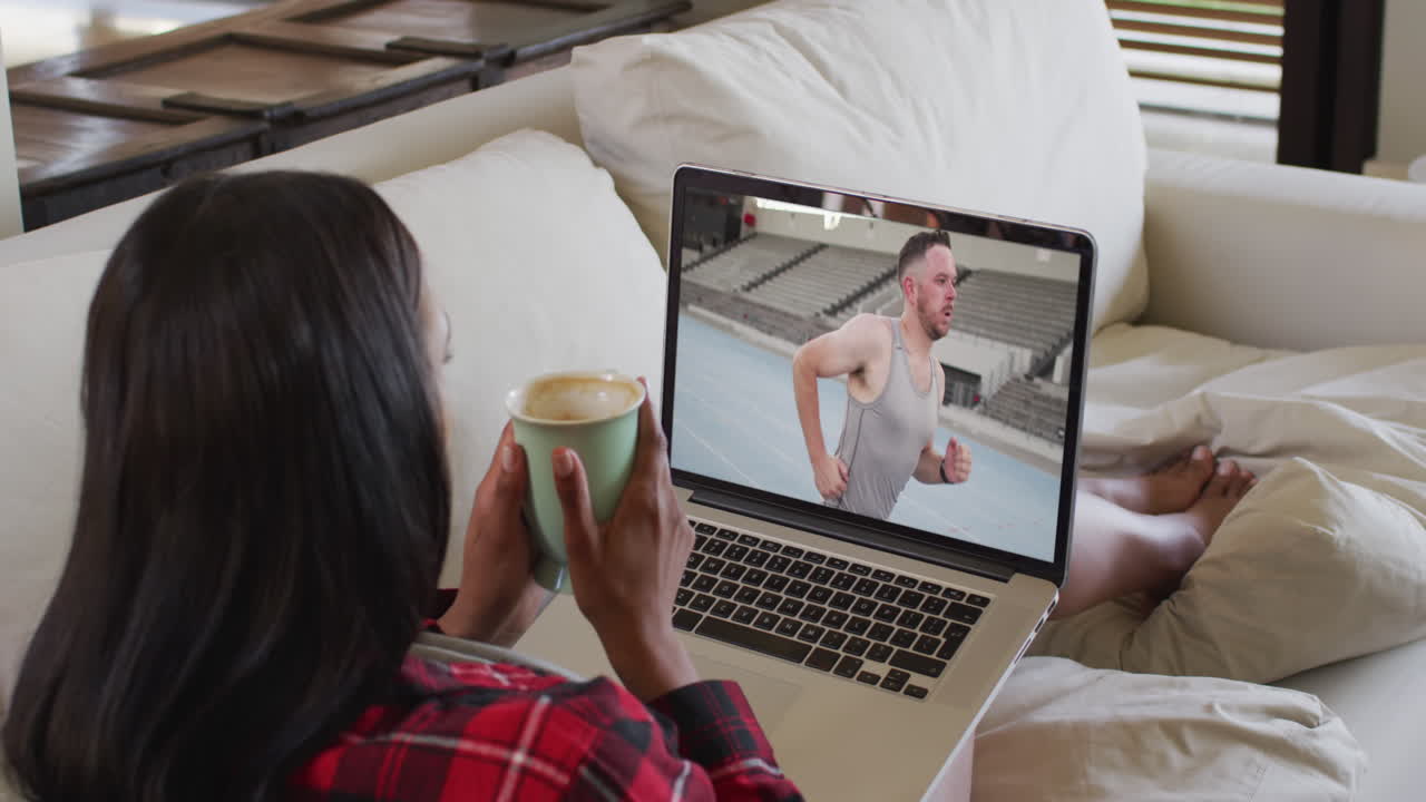 Composite of woman sitting at home holding coffee watching athletics event on laptop