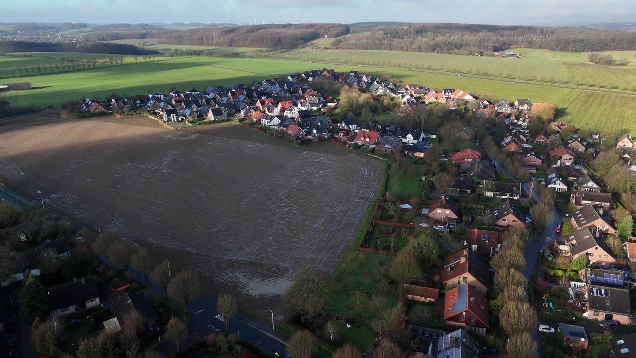 Aerial flyover muddy farm field in suburb neighborhood during sunny day. Single family homes with solar panels and rural hills in distance. Forward wide shot.