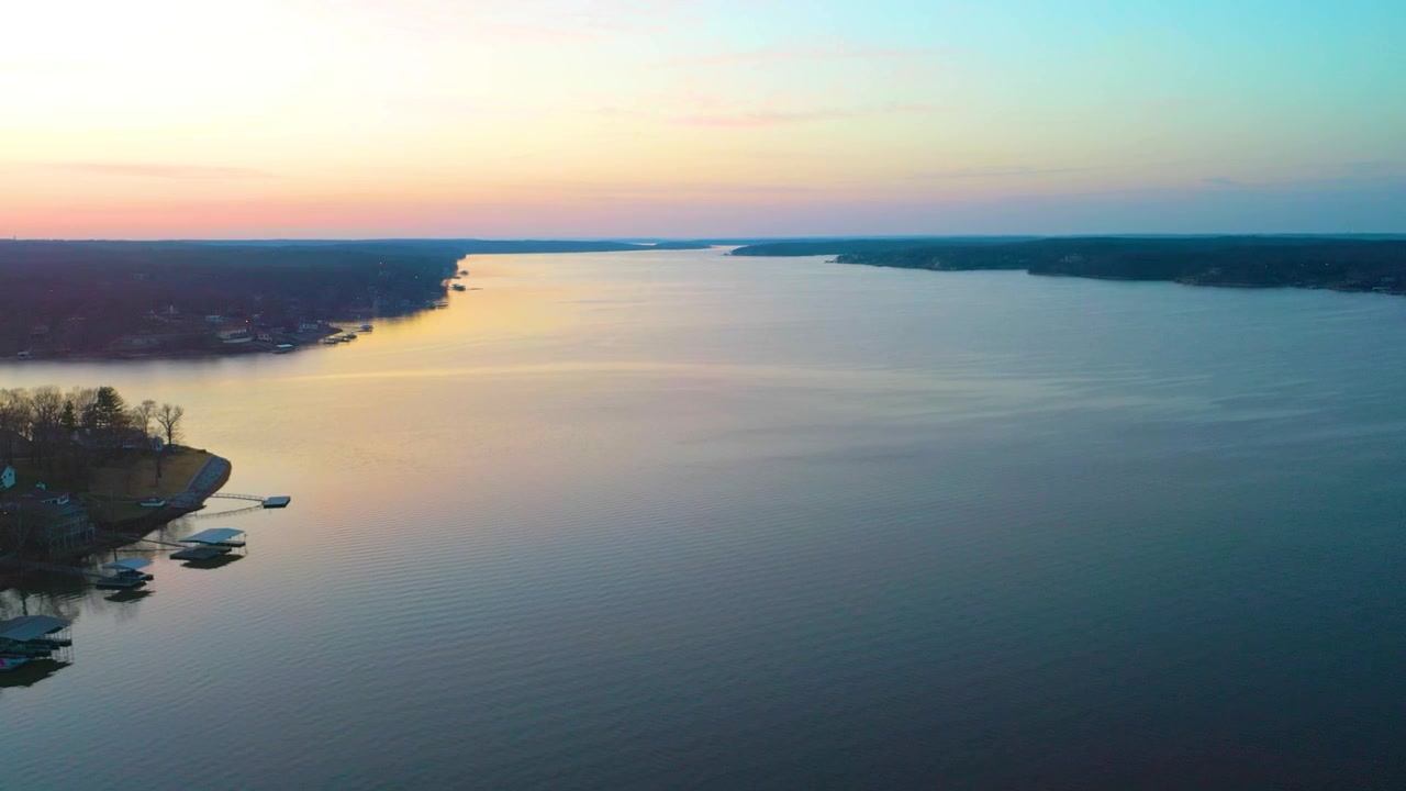 Záběry z dronu nad jezerem Beaver Lake v Arkansasu při západu slunce, ukazující klidnou modrou vodu, zalesněné pobřeží Ozarku, vzdálené domy, pastelovou večerní oblohu a teplé odrazy na hladině nádrže.