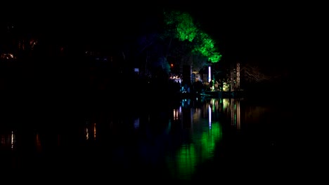 Multicolored-lights-at-night-live-concert,-singer-with-music-group-perform-near-city-park-lake