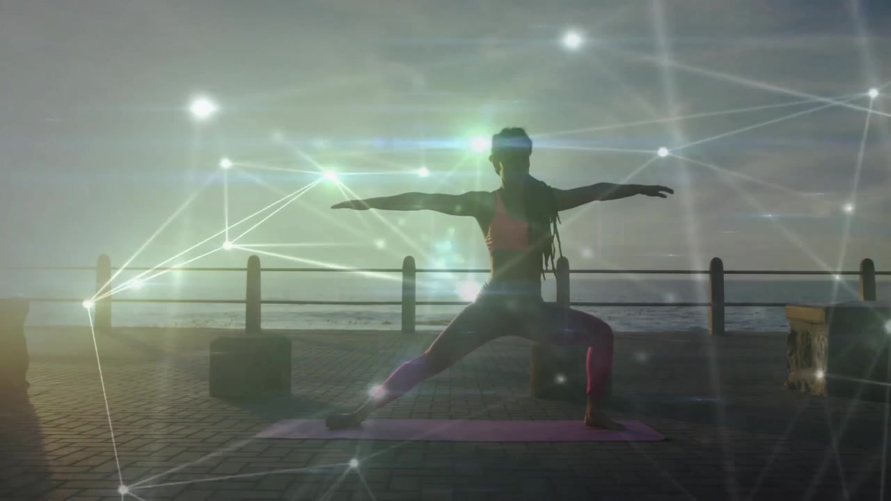 Mulher desenrolando tapete de yoga ao amanhecer à beira-mar, mantendo a pose do guerreiro II enquanto malha brilhante pulsa para bem-estar.