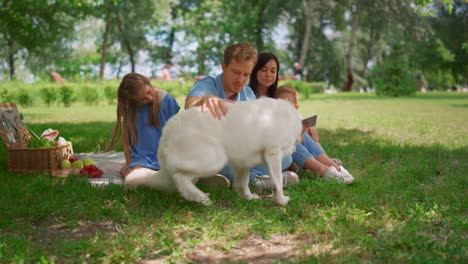 white dog lie down near family holding tablet on picnic. father caress labrador.