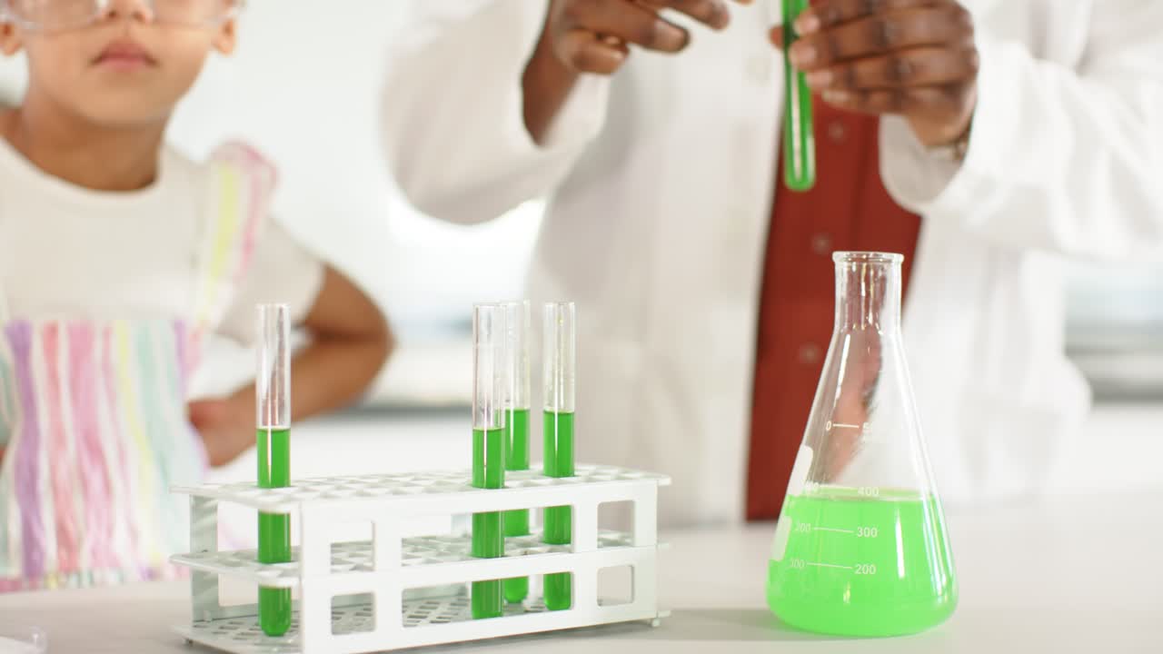 Female teacher showing to female youth at lab bench, reaching, arranging green test tubes in rack