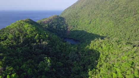 Aerial-flyover-lush-green-mountains-and-lake-in-El-Valle,-Samana-during-sunlight---Beautiful-idyllic-lake-surrounded-by-hills-and-Caribbean-sea-in-background