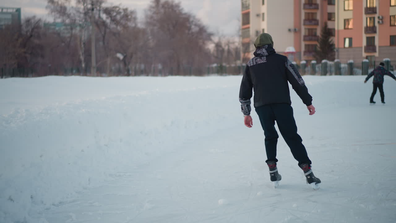 back view of adult skating on ice surface near learner practicing on frozen pond, ice scratched with skate marks, blurred snowy banks and urban buildings under soft winter light near trees