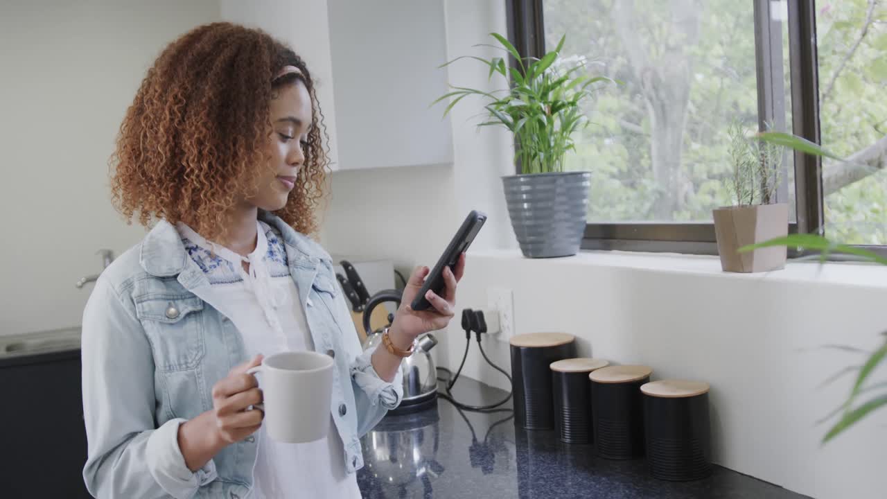 mujer biracial feliz usando un teléfono inteligente y bebiendo café en la cocina, cámara lenta