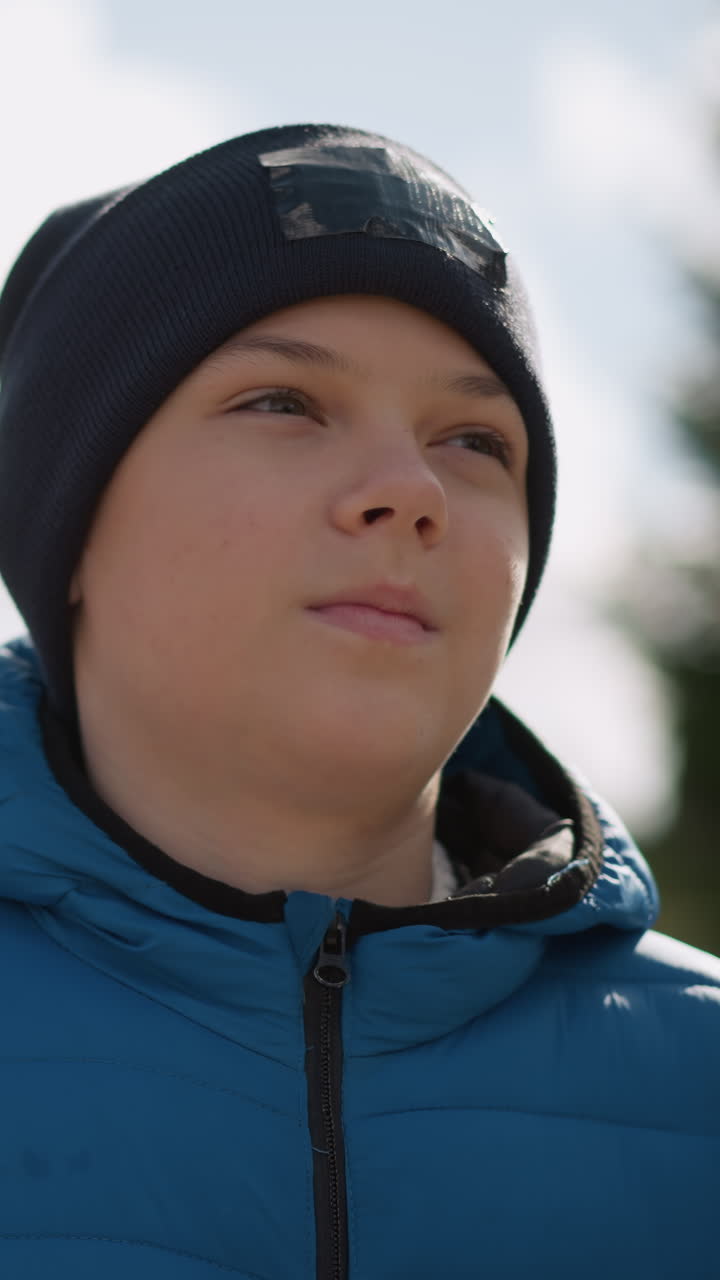 primer plano de un niño con chaqueta azul y gorra negra paseando por una zona residencial, mirando pensativo, con casas borrosas y árboles de otoño en el fondo