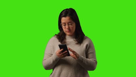 fotografía de estudio de una mujer joven usando un teléfono móvil para enviar mensajes de texto y desplazarse por internet de pie contra un fondo de pantalla verde
