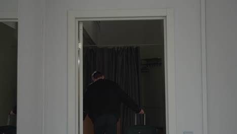 businessman wearing black jacket seen from behind as he enters room after business trip with curtain background and visible coat rack while gently closing white interior door