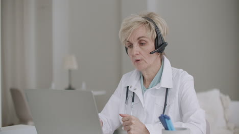 aged female physician is lecturing at online medical conference sitting in her office in hospital with laptop