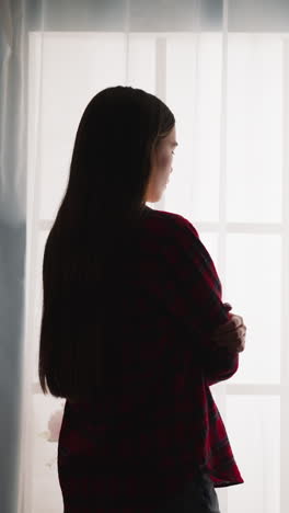 silhouette of woman with long hair in shirt stands near french window in room backside view slow motion. dreaming lady spends time alone at home