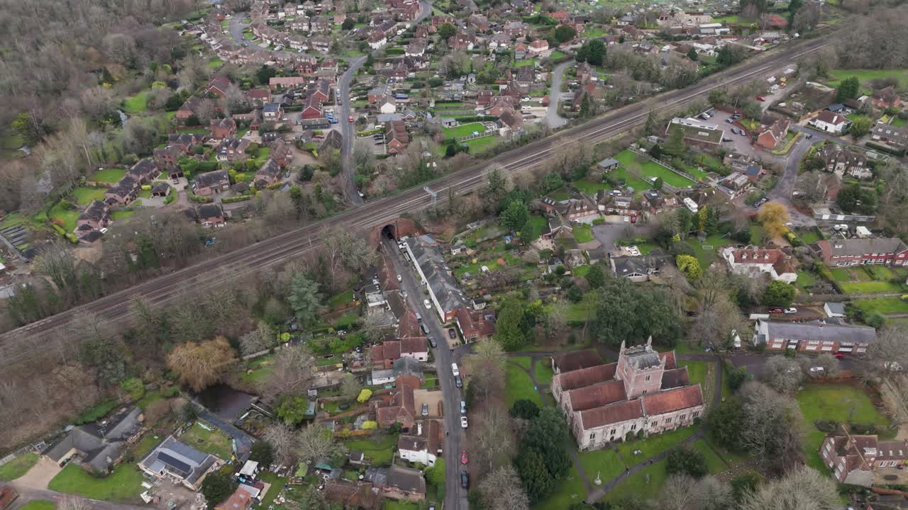 Aerial tracking view of countryside railway threading between tiled-roof cottages, gardens and woodland, capturing rural rail connectivity through an English village