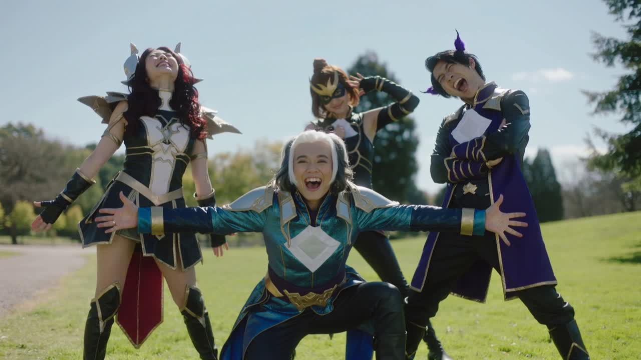 Güneşli bir parkta fantezi zırh içinde kutlama pozu veren dört cosplayer, omuzluklarını sergiliyor