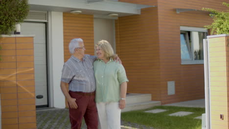 feliz pareja mayor de pie junto a la nueva casa, abrazándose y hablando, luego sonríen a la cámara