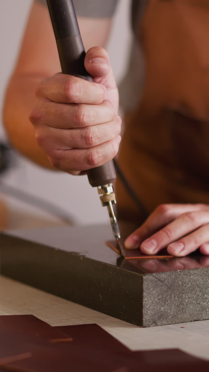 Leathersmith polishes leather piece edge with tool at equipped workplace closeup. Master works with traditional material in studio. Skilled handyman