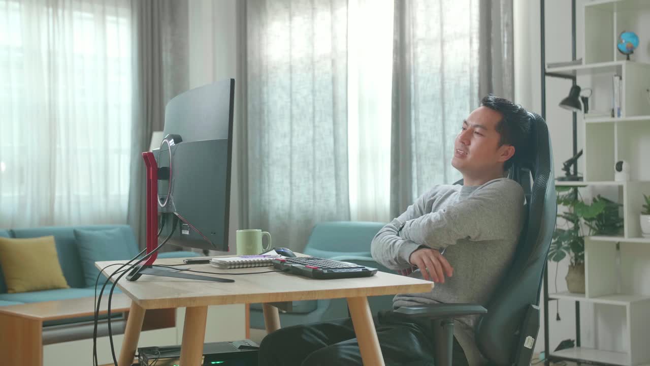 Asian Man In Long Sleeved T-Shirt And Black Pants Crossing His Arms And Leaning Back While Using Desktop Computer For Working At Home.