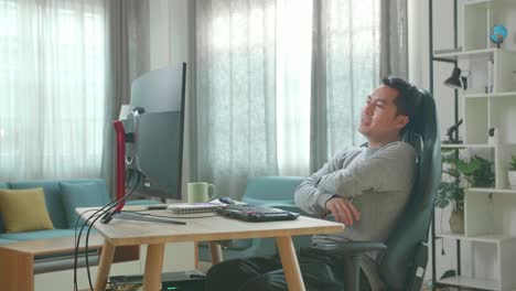 hombre asiático con camiseta de mangas largas y pantalones negros cruzando los brazos y inclinándose hacia atrás mientras usa la computadora de escritorio para trabajar en casa.