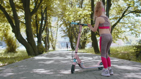 Una-Joven-Rubia-En-Forma-Atlética-Comienza-A-Viajar-En-Scooter-Eléctrico-En-La-Carretera-En-El-Parque-En-Un-Día-Soleado