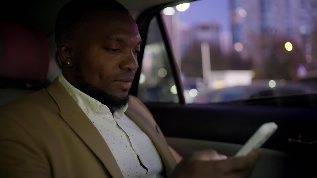 검은 피부와 짧은 헤어스타일과 갈색 슈트를 입은 수염을 가진 자신감있는 남자가, 사업가, 저녁 도시를 따라 운전하고 현대적인 자동차 인테리어에서 그의  스마트폰에서 일합니다.