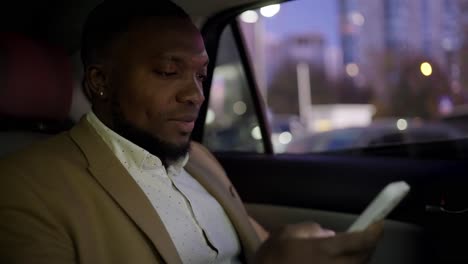 A confident man with Black skin and a short haircut with a beard in a brown suit, a businessman, drives along the evening city and works on his white smartphone in a modern car interior