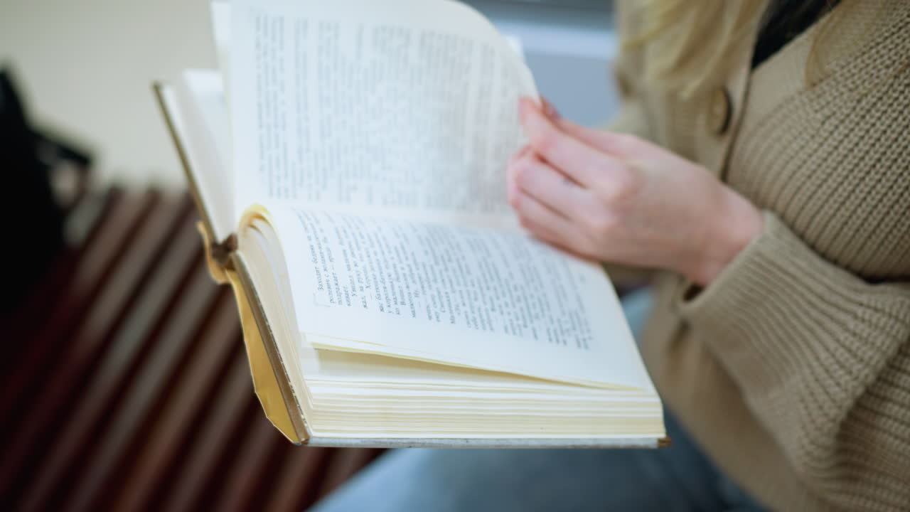 vista de cerca de la mano de la dama blanca girando a una nueva página, con pantalones de jean y suéter de diseño tejido, con la estructura moderna borrosa en el fondo