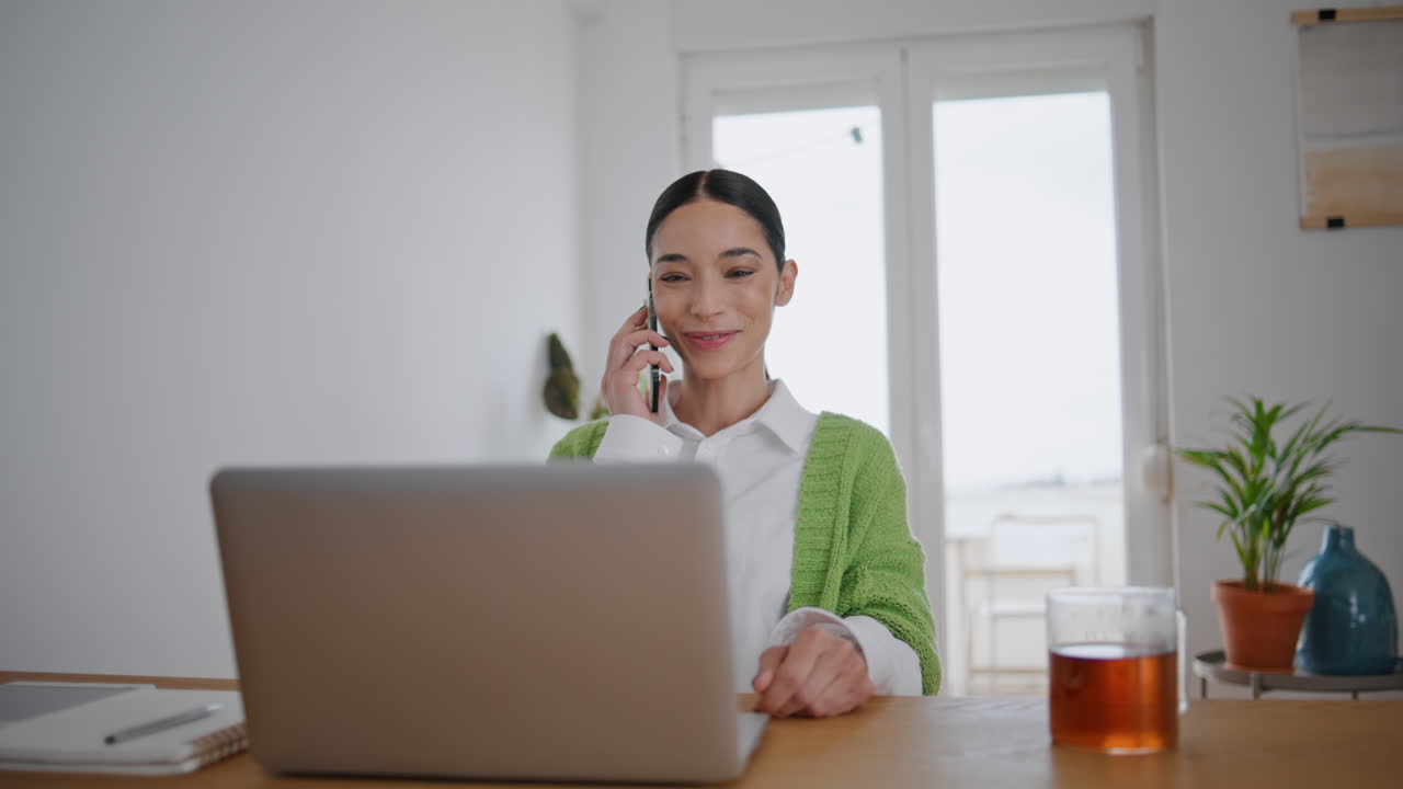 コンピューターのそばでスマートフォンを手に取る微笑むビジネスウーマンのクローズアップ。話す女の子。