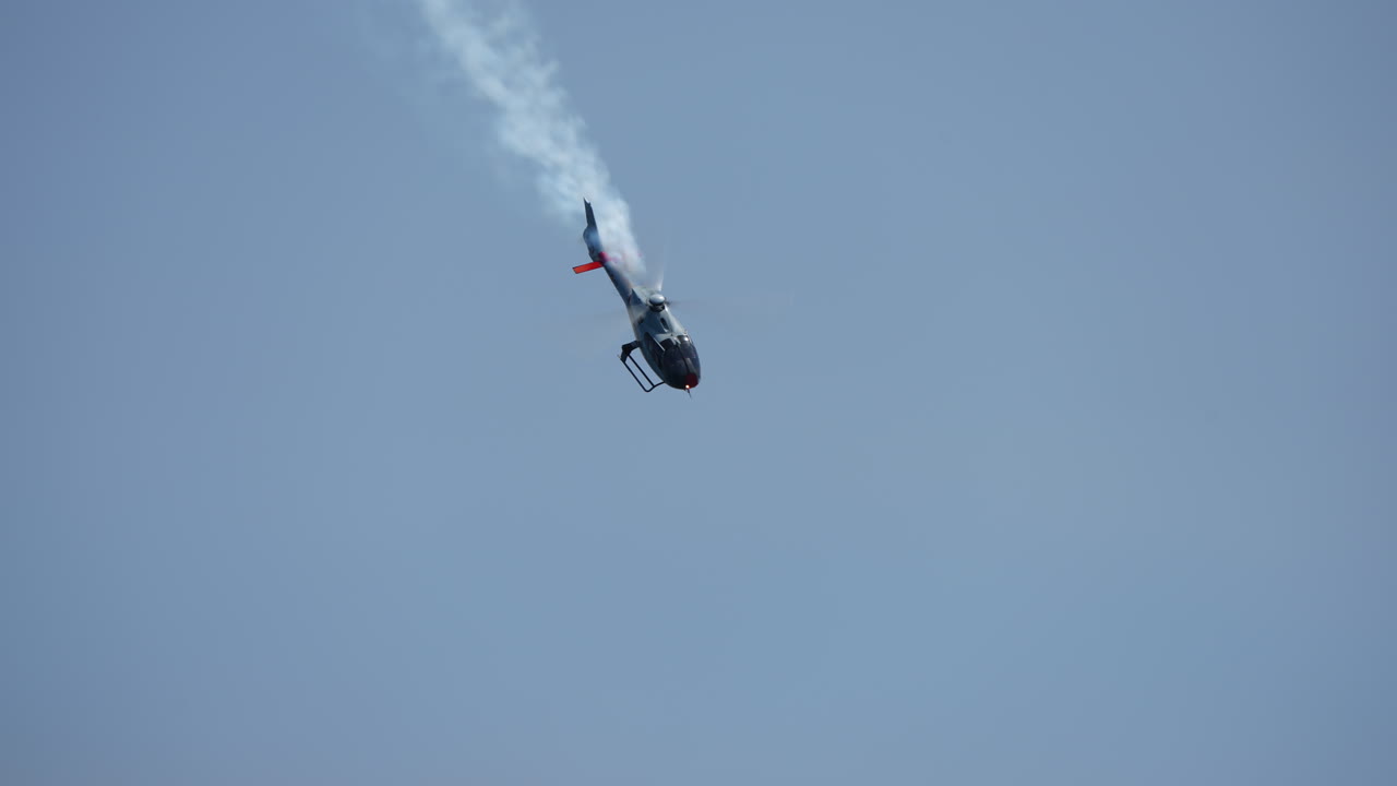 Aerobatic stunt helicopter executing a vertical flip in clear blue sky with visible smoke trail