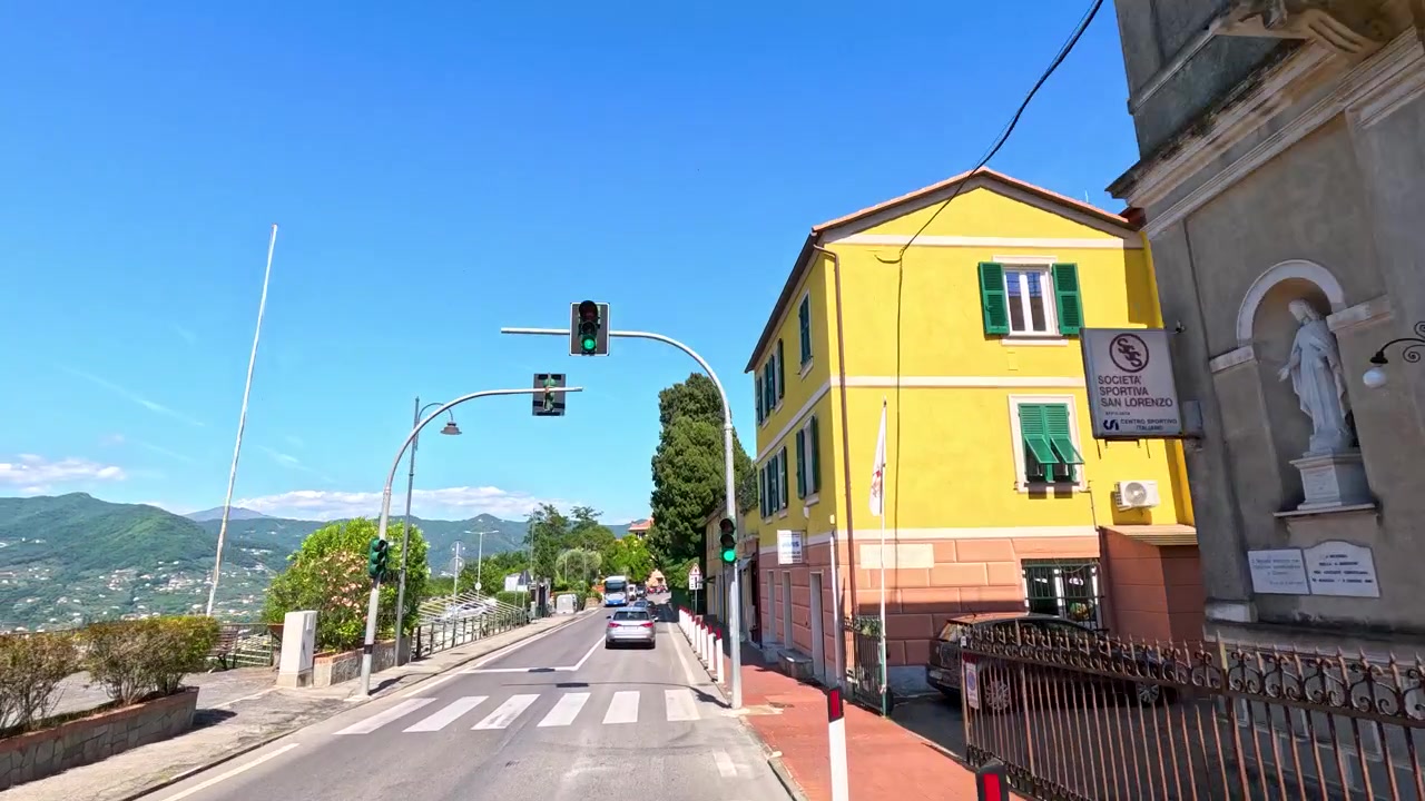 Um veículo trafega por uma rua ensolarada, passando por edifícios históricos e uma torre de igreja