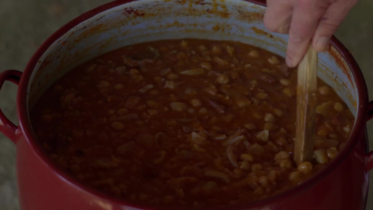 Una mano remueve un guiso de frijoles sustancioso a fuego lento en una olla rústica roja. Un primer plano resalta la textura, el color y el sabroso ambiente de cocina casera.