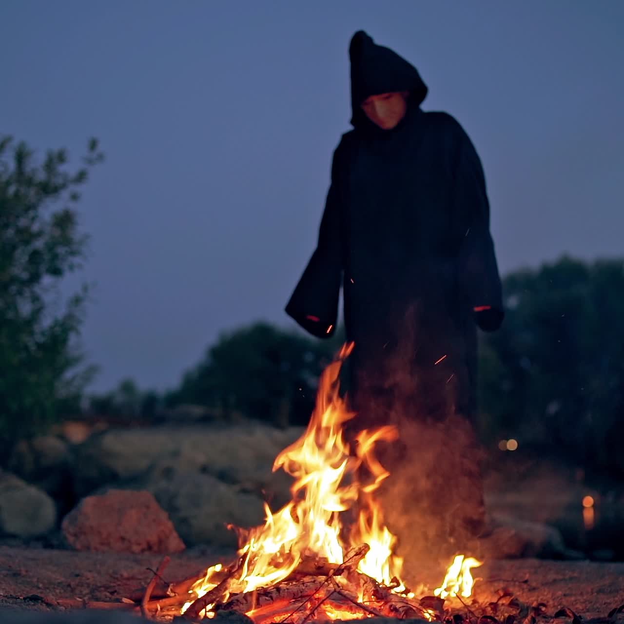 Witch evil in black gown near fire at night. Mysterious wizard doing shamanism over the flame. Scary figure in black making ritual. Halloween night.