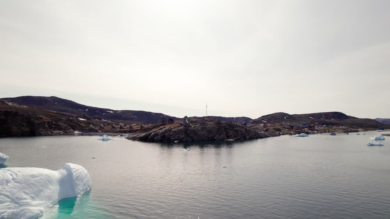 Rotating drone shot circling Ilimanaq settlement in Greenland, with massive icebergs drifting in the Arctic sea in front of the village