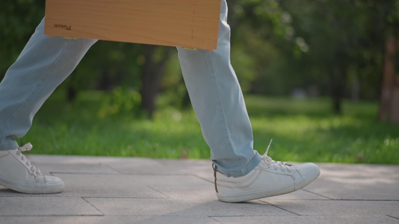 Kafkas bir sanatçının kaldırımda yürümesi, spor ayakkabılarına ve ahşap sanat kutusuna yakın çekim, denim pantolon, yeşil parkın yanındaki beton yolda ritmik yürüyüş, açık havada taslak çizmeye hazır, günlük modern hava