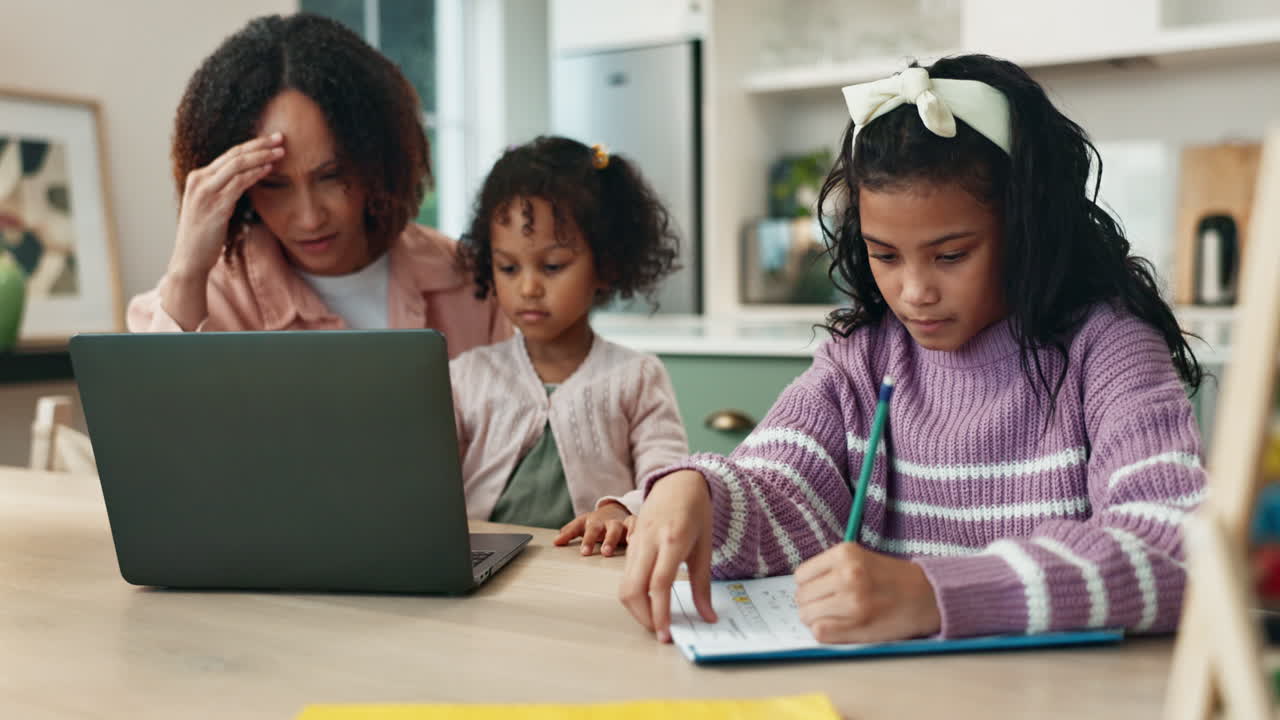 A woman looks stressed while working on her laptop while her two daughters are studying at the kitchen table.