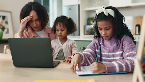 a woman looks stressed while working on her laptop while her two daughters are studying at the kitchen table.