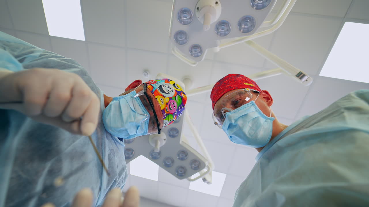Two male surgeons in funny caps applying tools at surgery. Low angle view at the confident doctors at the backdrop of lamps in surgical room.