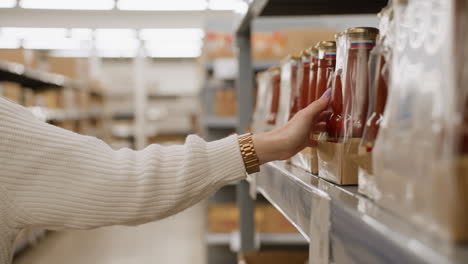 close up of browser hand touching glass tomato ketchup bottles lined on supermarket shelf rails in bright grocery aisle highlighting polished nails wrist watch and blurred background