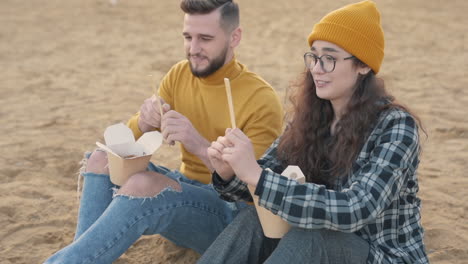 jóvenes amigos hombres y mujeres comiendo comida para llevar sentados en la playa