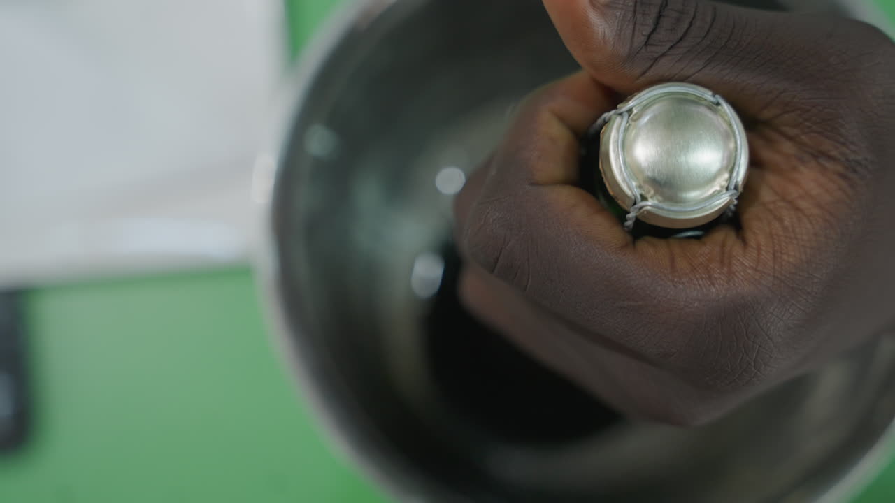 Mão negra destampa garrafa de champanhe, close-up de encontro mostrando mão abaixando garrafa verde em balde de gelo de metal, condensação e restos de folha metálica ao redor do gargalo, rolha removida, preparação romântica íntima.