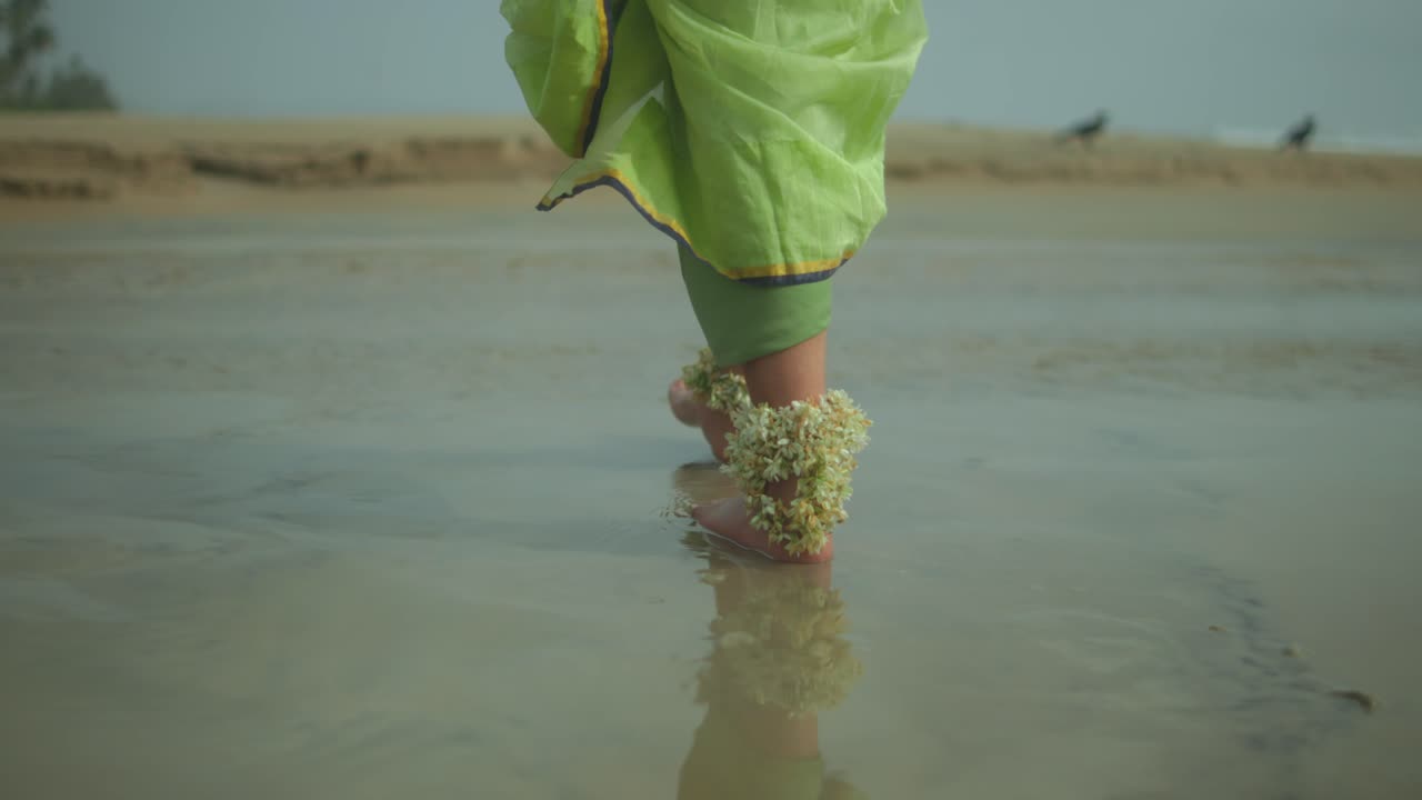 Primo piano di una donna a piedi nudi che indossa un sari verde e cavigliere floreali, che cammina lentamente sulla sabbia bagnata di una spiaggia tranquilla.