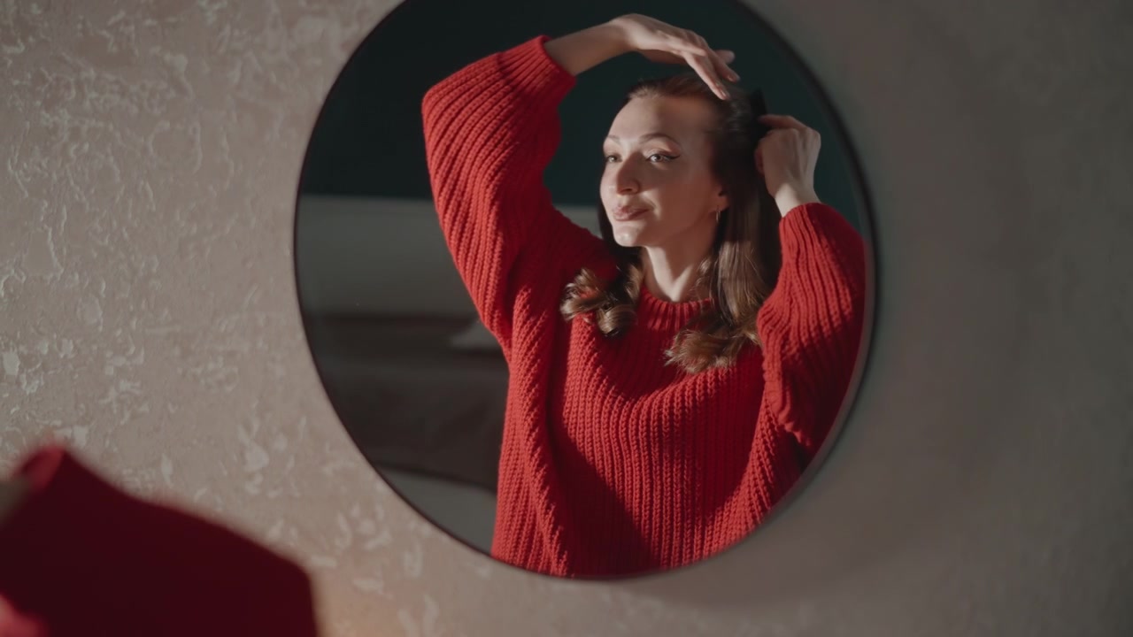 Mujer vista a través de un espejo de pared redondo se ajusta el cabello con las manos cerca de la frente mientras usa un suéter de punto rojo, iluminación lateral suave, fondo interior neutro, expresión relajada, ambiente tranquilo de autocuidado