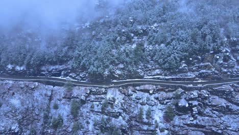 aerial footage of a snowy, scenic byway, winding mountain valley road during a snowstorm with pine trees, a mountain highway, rocky cliffs and forest during winter on a cold, blue day