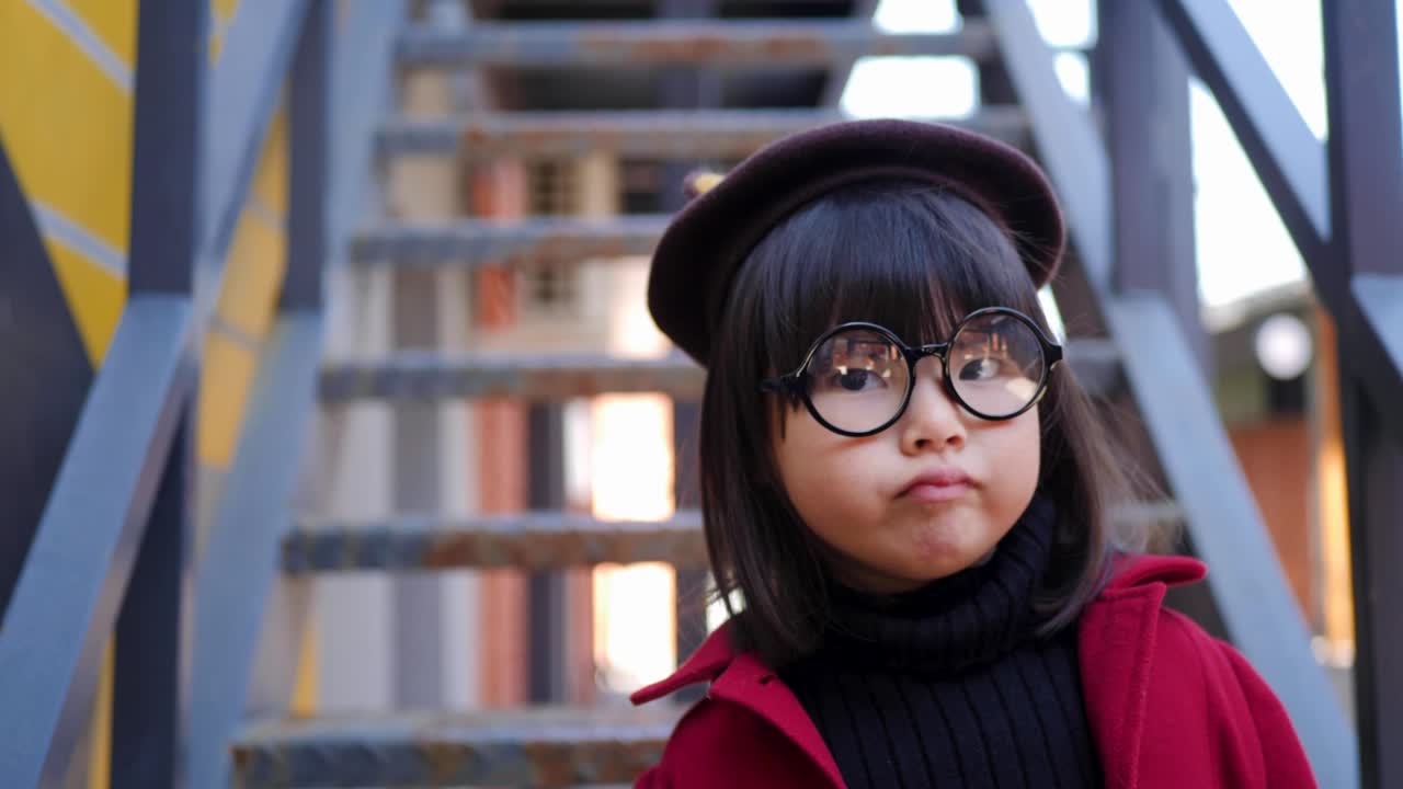 은 코트와 모자, 둥근 안경을 입은 한국 소녀가 가을에 철 계단에 앉아 있다.
