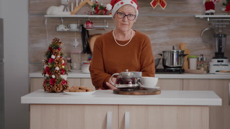 retrato de una mujer mayor con sombrero de santa de pie en la mesa en la cocina decorada con navidad