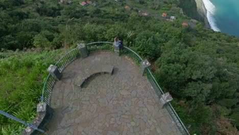 Una-Mujer-Sentada-En-Una-Valla-Con-Vistas-Panorámicas-Al-Océano-Y-A-La-Ciudad-De-La-Costa-De-Madeira