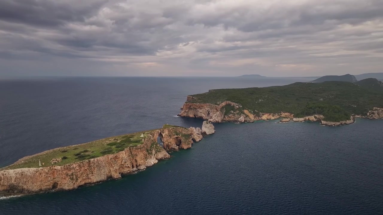Ripresa aerea ad alta quota con drone che mostra l'intera cresta dell'isolotto di Tsichli Baba con il suo arco marino naturale, allineato con l'isola di Sfacteria sotto un cielo crepuscolare nuvoloso vicino a Pylos, Grecia