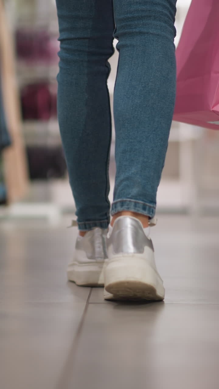 Leg view of shopper walking through retail clothing store aisle carrying colorful shopping bags in vibrant modern environment with neatly arranged racks