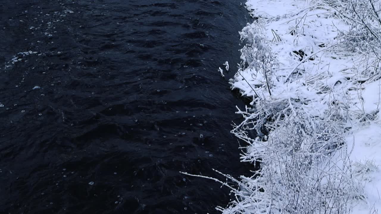 Dark cold and foamy river water flowing in slow motion at winter time while riverbanks are covered with white fluffy snow. Footage rises up and reveals more of the river and nature surrounding it.