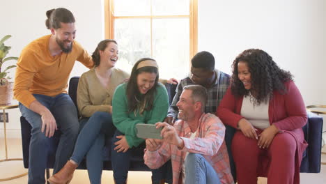 Happy-diverse-male-and-female-friends-relaxing-at-home-together-talking-and-looking-at-smartphone